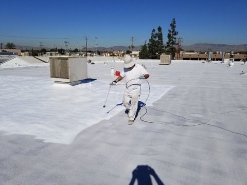 Worker in a white sun hat spraying bright white coating onto a flat commercial roof beside an HVAC unit.