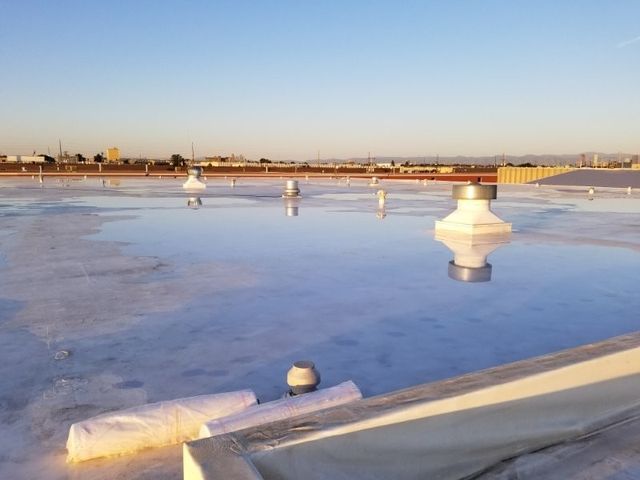 Flat commercial roof with standing water reflecting the sky, roof vents, and rolled materials at the edge.
