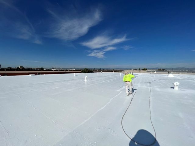 Worker in a neon safety shirt spraying white coating across a finished flat commercial roof.