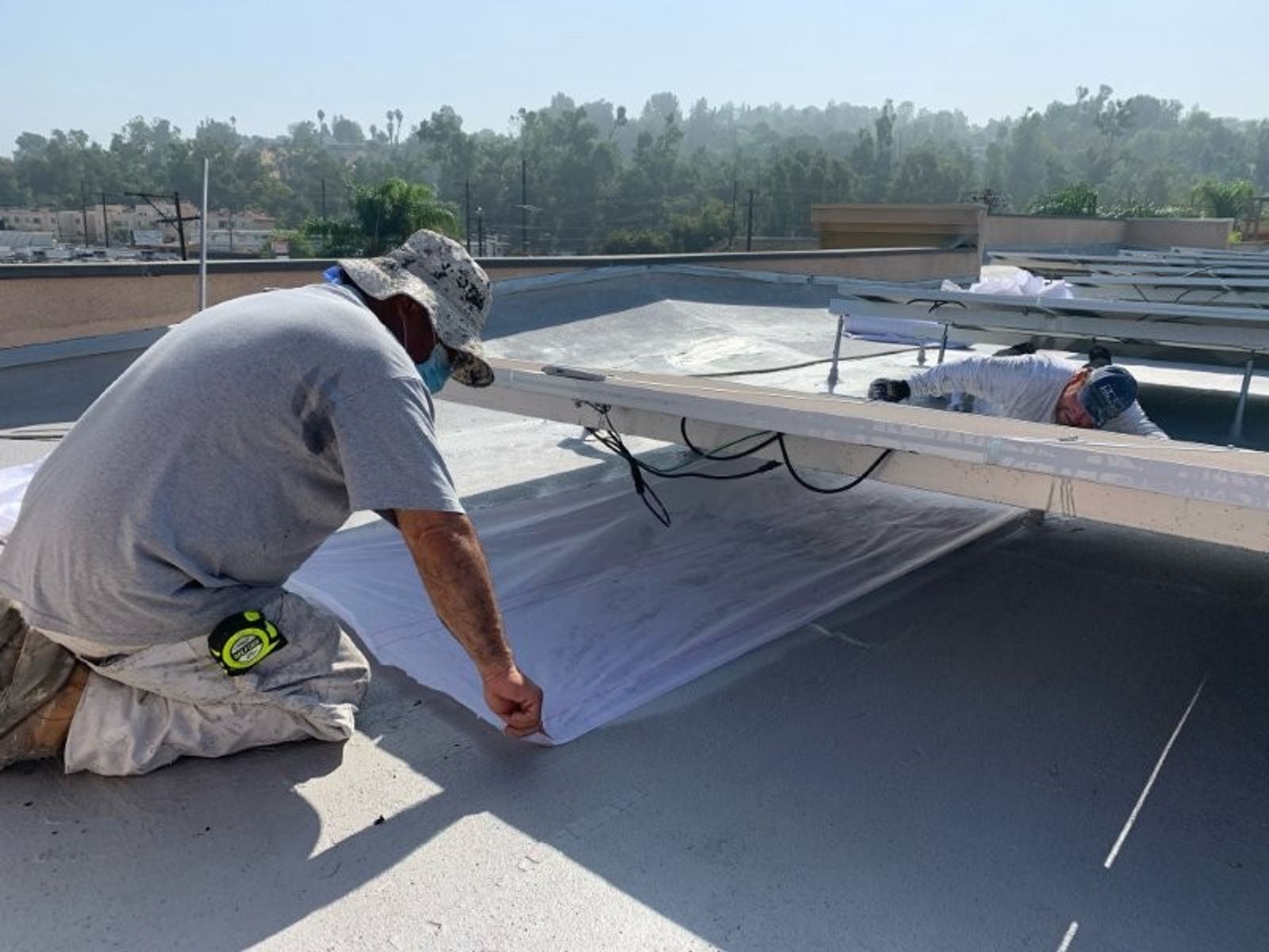Two workers on a flat commercial roof positioning white sheet material around metal racking supports under a hillside skyline.