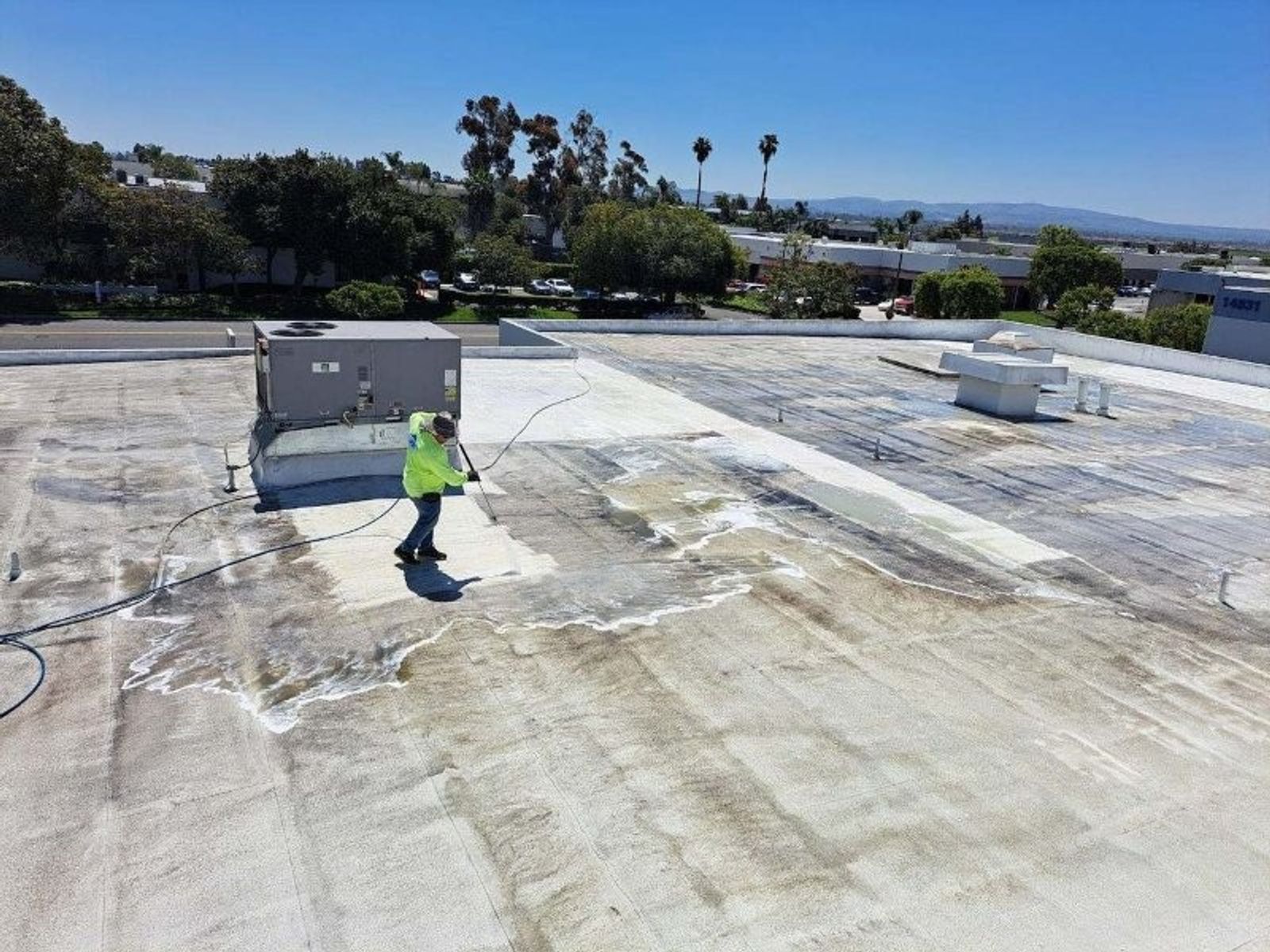 Worker in a neon safety shirt pressure washing a flat commercial roof, with cleaned light areas next to dark soiled membrane.